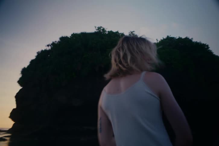 A blonde woman turns to face the camera in slow motion on a dark, moody beach as her hair blows in the wind at dusk