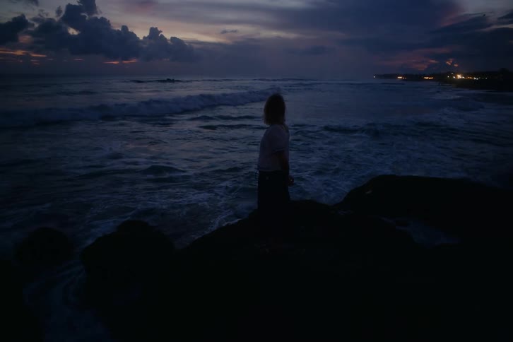 A silhouetted woman stands on a rock contemplating the ocean waves crashing against the shore at twilight