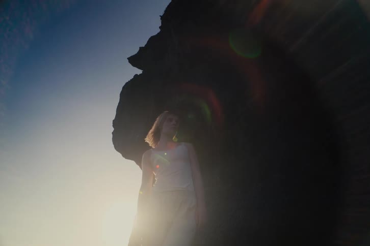 Cinematic slow-motion shot of a contemplative young person standing against a silhouetted rock formation as the sun flares behind them