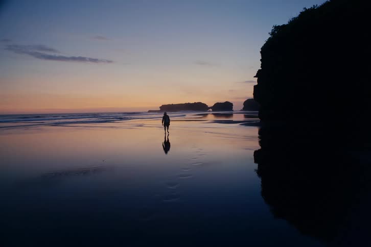 The silhouette of a person walks away on a tranquil beach at twilight, their reflection mirrored on the wet sand