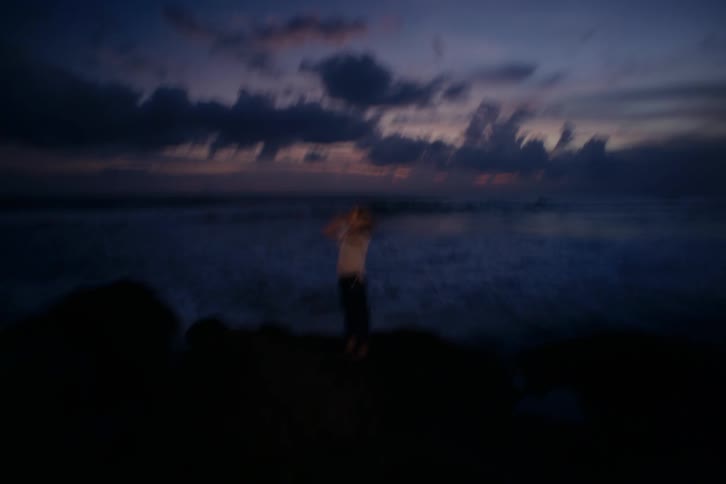 A dreamy, abstract shot captures a woman standing on rocks, watching the ocean waves roll in at twilight