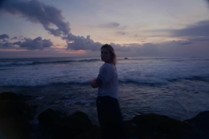 Dreamy long exposure shot of a beautiful young woman on a rocky beach at twilight, turning to look into the camera