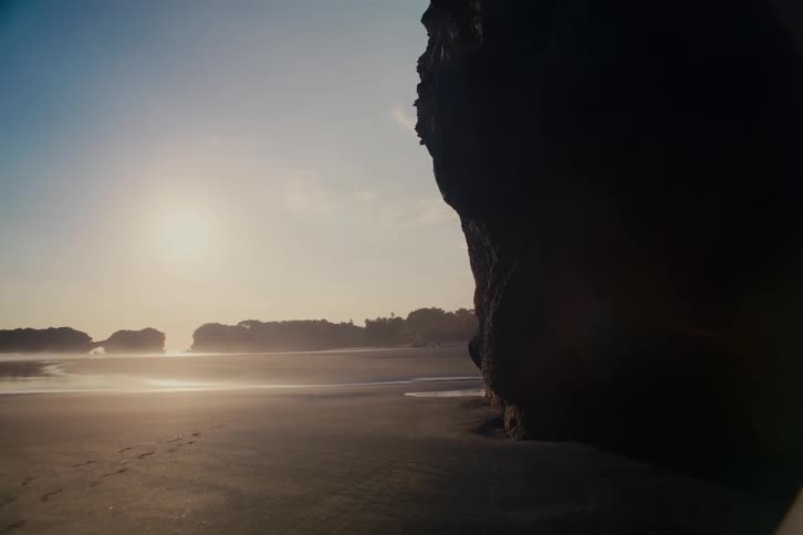 The bright sun hangs low over a serene, empty beach at low tide, illuminating footprints in the dark sand next to a silhouetted rock formation