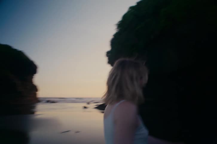 A carefree young woman with blonde hair runs and smiles on a remote beach at dusk in slow motion