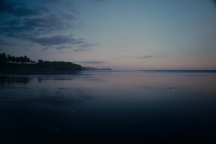A serene view of a dark, tropical beach at twilight where the calm ocean perfectly reflects the moody sky