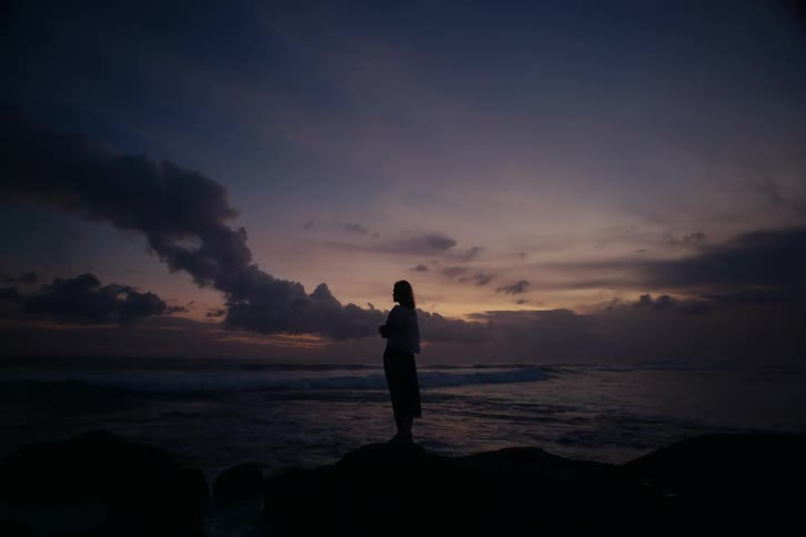 A silhouetted woman stands peacefully on rocks by the ocean, watching the dramatic purple and orange sky at dusk