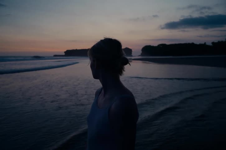 A silhouetted woman stands on a quiet beach at dusk, looking out at the ocean as the last light of sunset fades