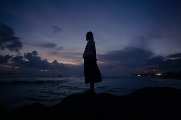 A pensive woman in silhouette stands on a coastal rock, gazing at the ocean waves during a dramatic twilight