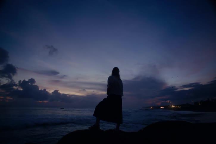 A silhouetted woman stands contemplatively on a coastal rock, gazing at the ocean waves under a dramatic twilight sky