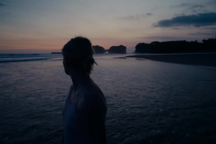 A silhouetted woman stands peacefully on the shore, looking out at the ocean waves as dusk settles on the horizon