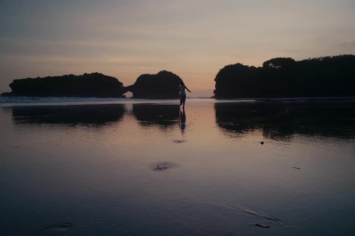 A silhouetted woman gracefully runs away from the camera in slow motion on a serene, reflective beach at twilight