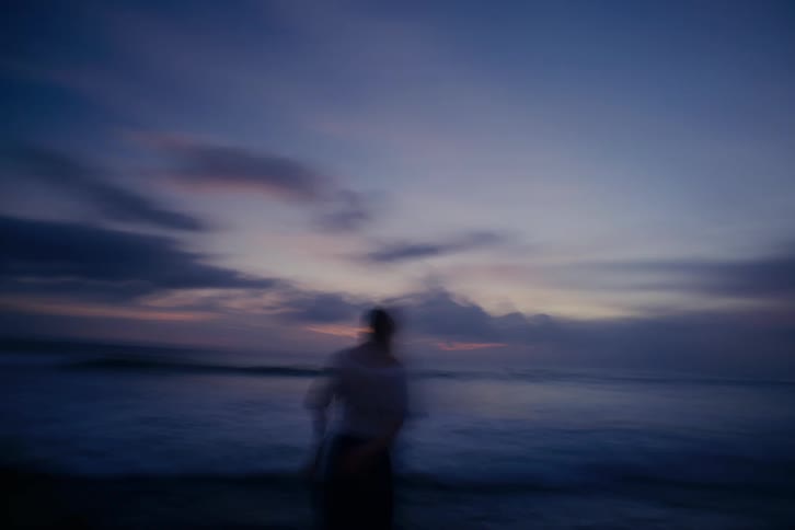 An abstract, dreamy, and blurry handheld shot reveals a young woman standing on the shore at twilight, looking back at the camera
