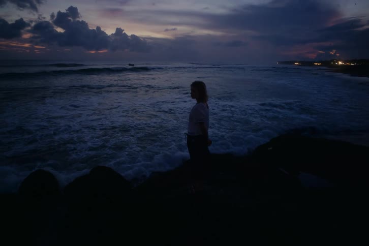 A solitary woman stands silhouetted on a rocky shore at dusk, contemplating the crashing waves of the ocean
