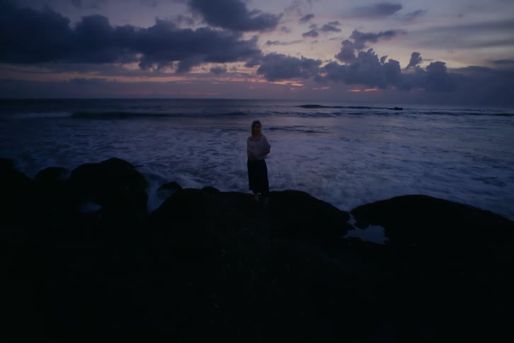 A woman stands still on the rocks watching the ocean waves at twilight in a long exposure shot