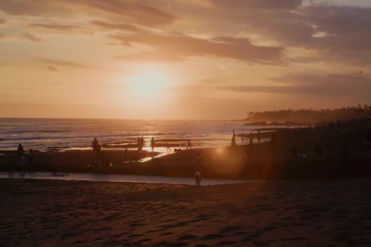 A crowd of people enjoy a beautiful golden sunset on a tropical beach, their silhouettes dotting the shoreline