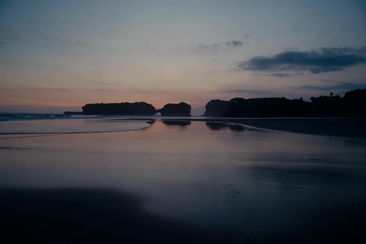 A peaceful and tranquil ocean shoreline at twilight reflects the silhouetted rock formations against the colorful sky