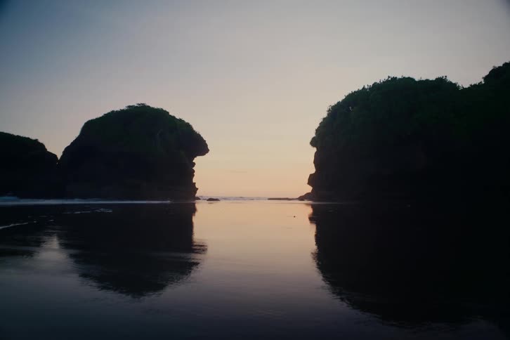 A serene view of two silhouetted rock formations reflected in the calm ocean water during a peaceful sunset