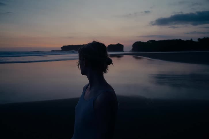 Cinemagraph shows a woman in silhouette standing on a beach at dusk, peacefully watching the ocean waves