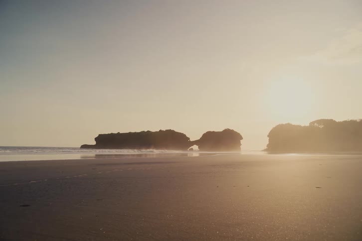 The sun sets over a tranquil, sandy beach, casting a golden haze over the calm ocean waves and distant rock formations