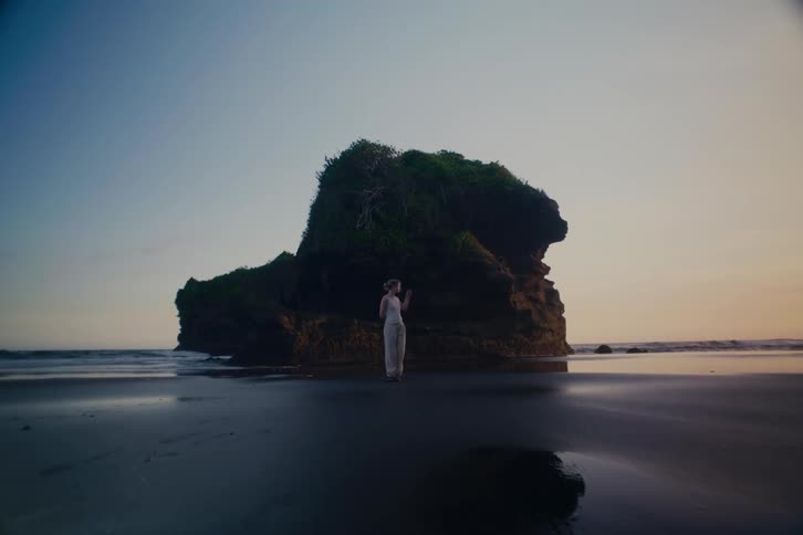 A lone woman stands on a dark sand beach, dwarfed by a massive rock formation as the sun sets over the ocean