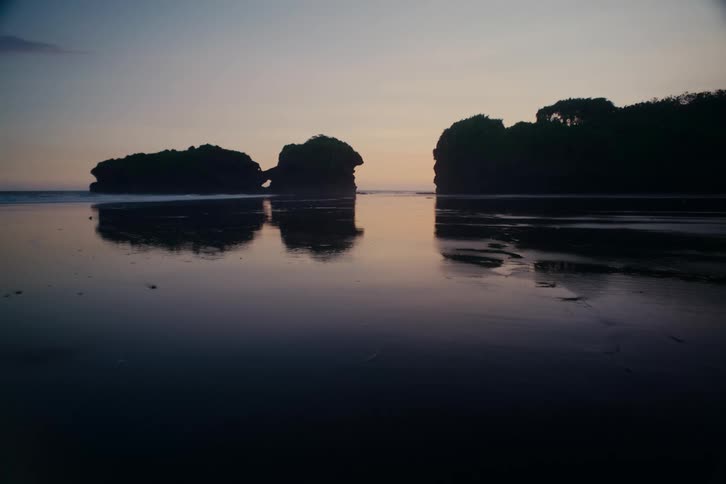 A tranquil and moody view of a silhouetted coastline with rock formations reflected in the wet sand at dusk