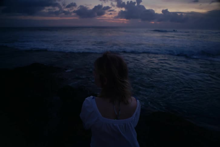 A lone woman sits on a rocky shoreline watching the ocean waves crash as the last light of day fades into night