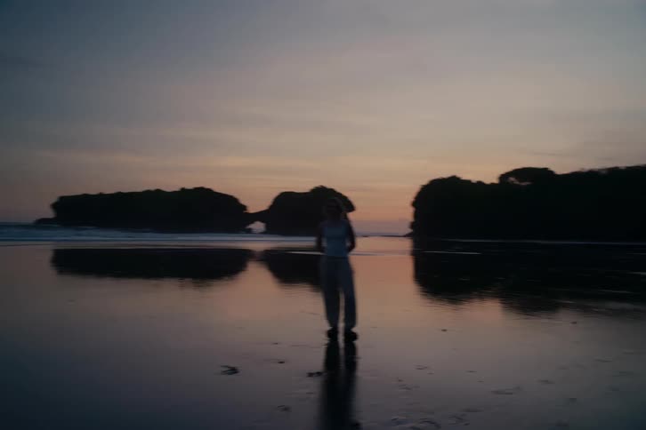A woman stands silhouetted on a reflective beach as the camera slowly pulls back during a tranquil sunset