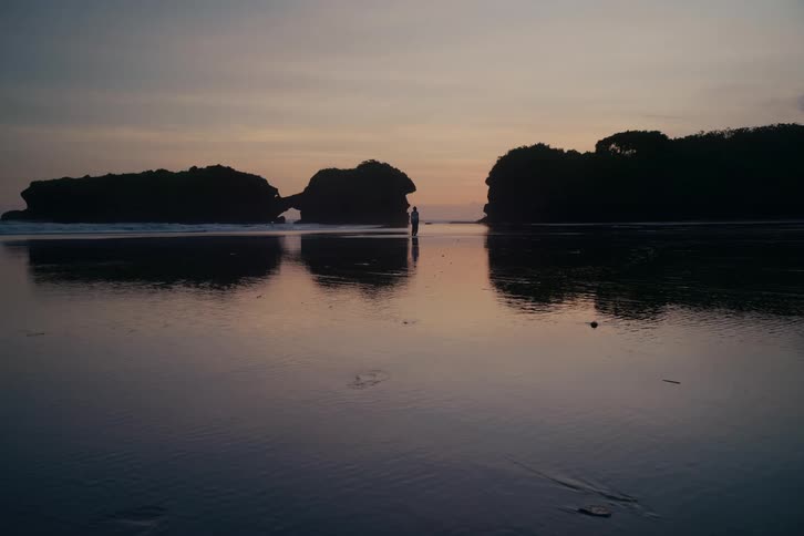 A lone figure stands silhouetted on a tranquil beach at dusk, with the calm water reflecting the sky and distant rock formations