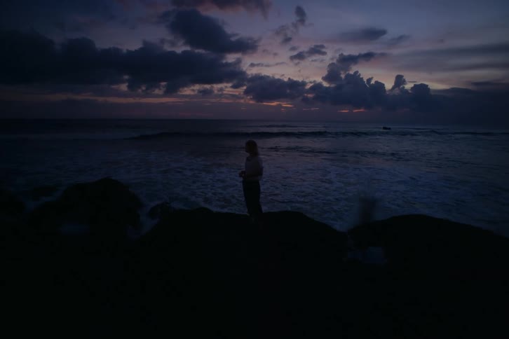 A silhouetted person stands on a rocky shore watching the ocean waves under a moody twilight sky