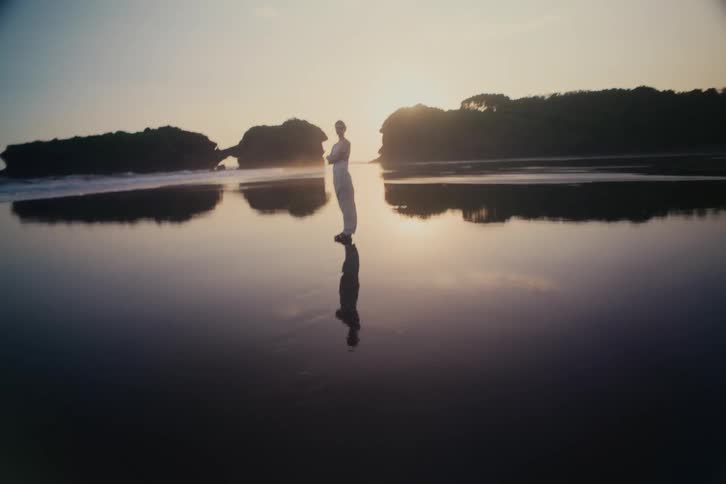 A serene woman stands on a wet, reflective beach at golden hour, with rock formations in the background, as the camera slowly zooms in for a close-up