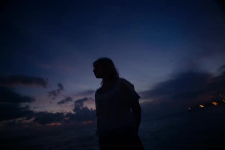 The dark silhouette of a woman looks out over the ocean against a deep blue twilight sky