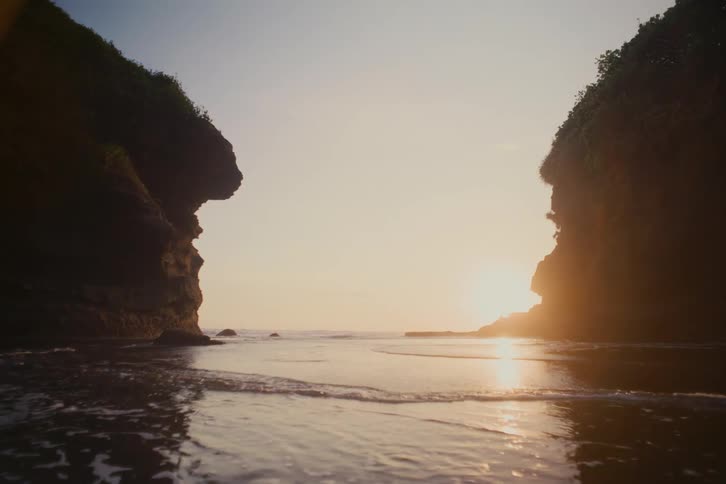 Calm ocean waves wash ashore between two large cliffs on a sandy beach during a golden sunset