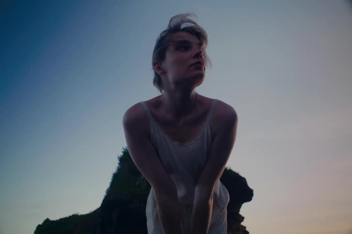 A pensive young woman in a white dress stands silhouetted against a dusky sky on a rocky coast
