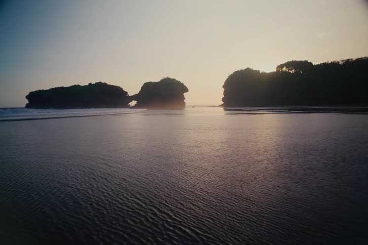A tranquil beach scene shows calm waters and distant, silhouetted rock formations with a natural arch during a golden sunset