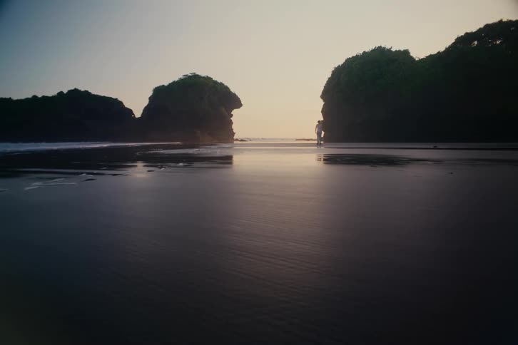A loving couple dressed in white walks hand-in-hand along a wide, reflective black sand beach at sunset