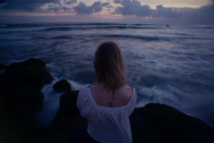 A woman seen from behind sits peacefully on dark rocks, watching the ocean's rolling waves at twilight