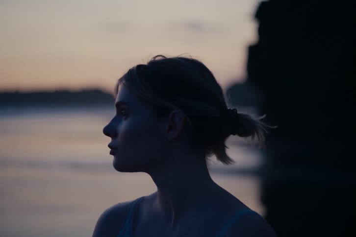 A contemplative young woman, silhouetted against the water at twilight, slowly turns to look directly at the camera