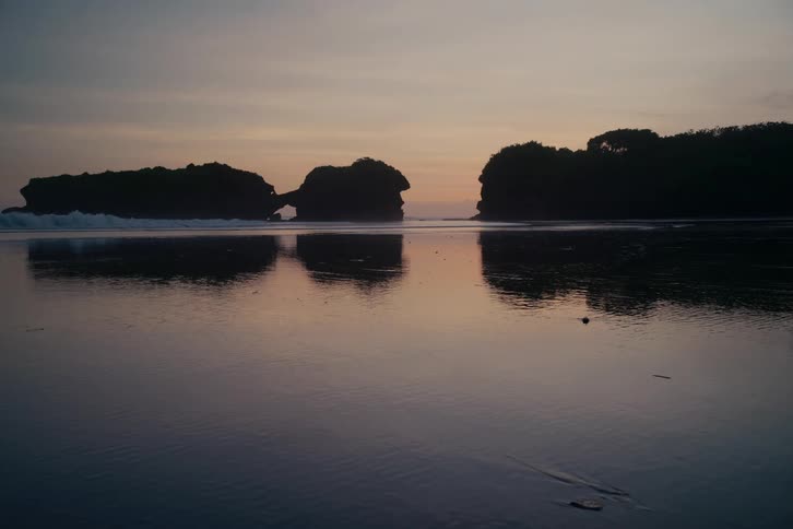 A tranquil beach scene at dusk shows silhouetted rock formations with a natural arch reflected in the calm, receding tide