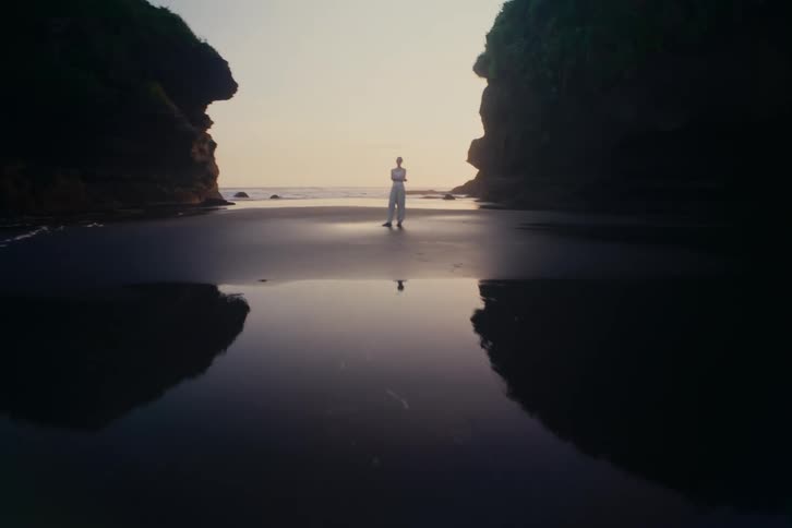A slow push-in on a young woman standing with arms crossed on a dark reflective sand beach between two cliffs at sunset