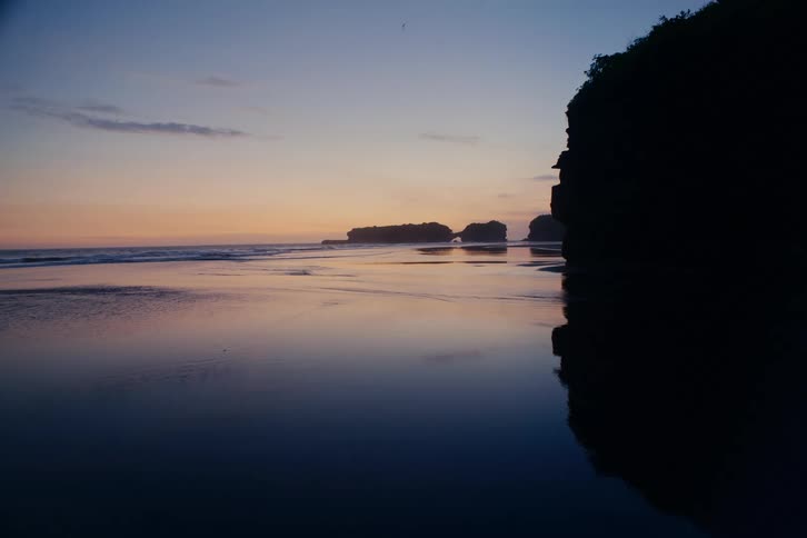 A tranquil seascape at twilight with calm, reflective water mirroring the colorful sky and silhouetted cliffs