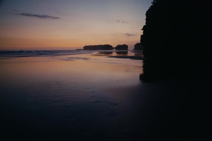 The tranquil ocean reflects the colorful twilight sky on a secluded beach with silhouetted rock formations and footprints in the sand
