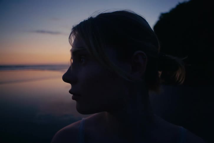 A thoughtful young woman stands silhouetted on a beach at dusk, turning her head from the ocean to look directly into the camera