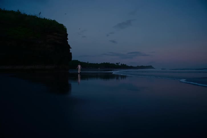 A woman in a white dress walks along the shoreline of a dark sand beach at dusk, with her reflection visible on the wet sand