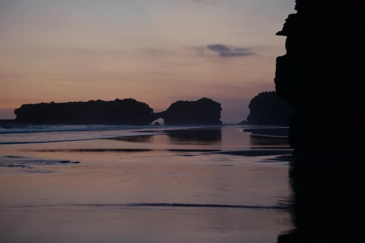 A tranquil beach at twilight with gentle waves and silhouetted rock formations reflected in the wet sand
