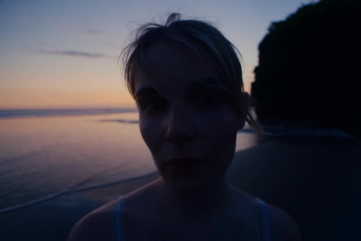 A moody, atmospheric close-up shows a young woman's face in silhouette as she looks at the camera and then turns to watch the ocean at twilight