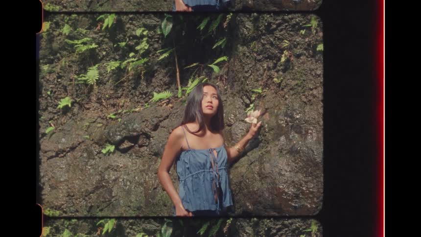 A contemplative woman in a blue top holds a flower and gently touches a wet rock face in a lush jungle, captured on vintage-style film