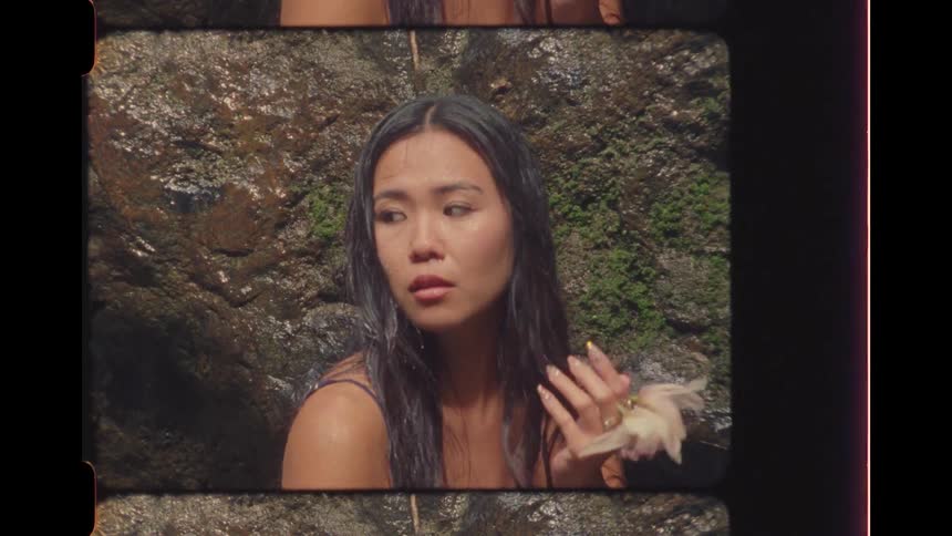 A serene Asian woman with wet hair holds an orchid flower by a waterfall, filmed in a vintage Super 8 style