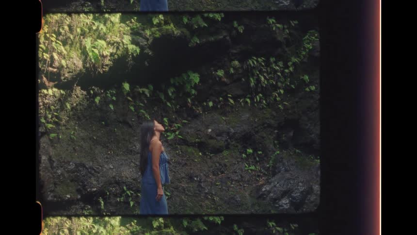 A woman in a blue dress looks up in wonder while standing in a lush, rocky jungle, captured on vintage 16mm film