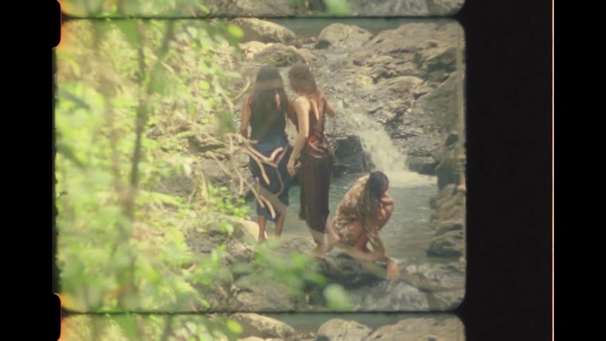 Vintage 8mm style footage shows three women in long dresses relaxing by a serene jungle stream and waterfall
