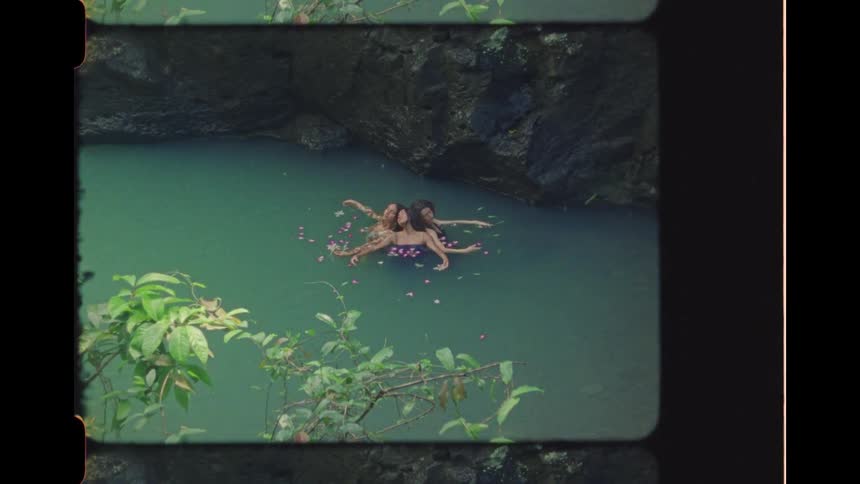Retro-style high angle footage shows three women floating together in a serene turquoise river surrounded by pink flower petals